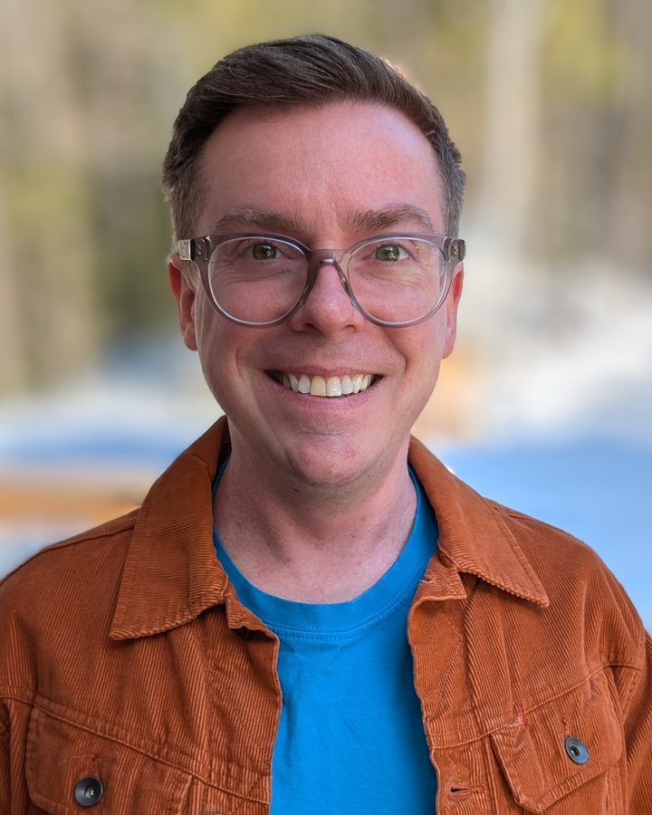 Dustin Faraco, smiling, wearing glasses and an orange corduroy jacket